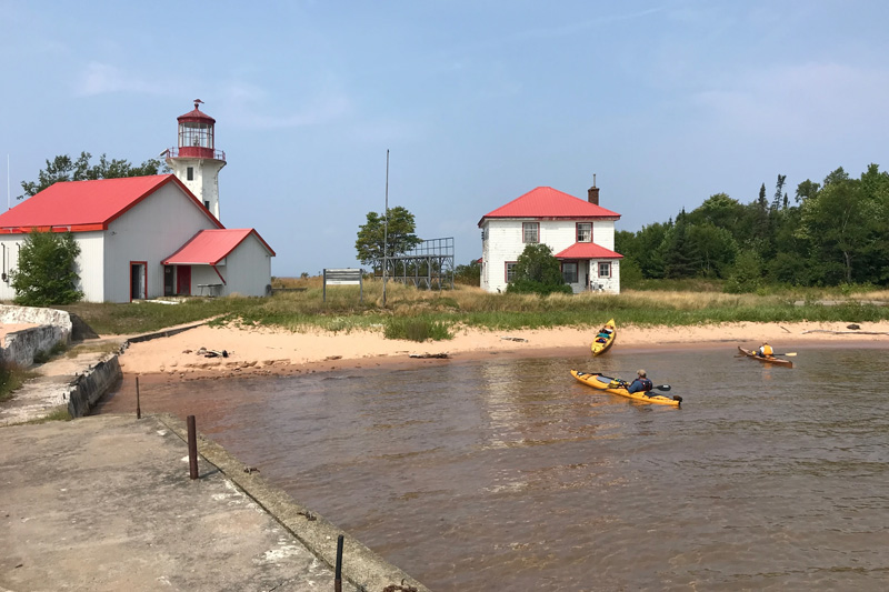 kayakers approaching a lighthouse station and old house, both white with red roofs, with a sandy beach