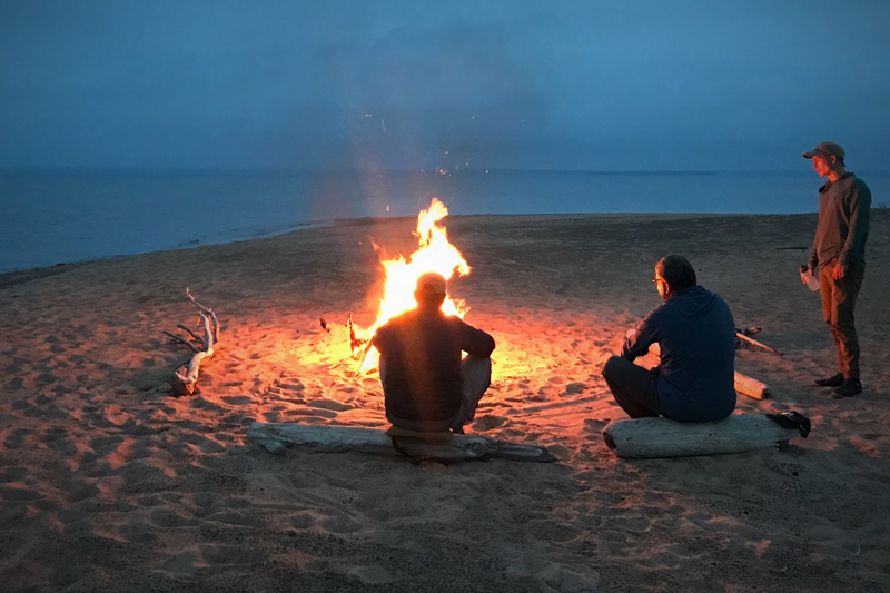 three people relaxing on driftwood logs around a campfire on the beach